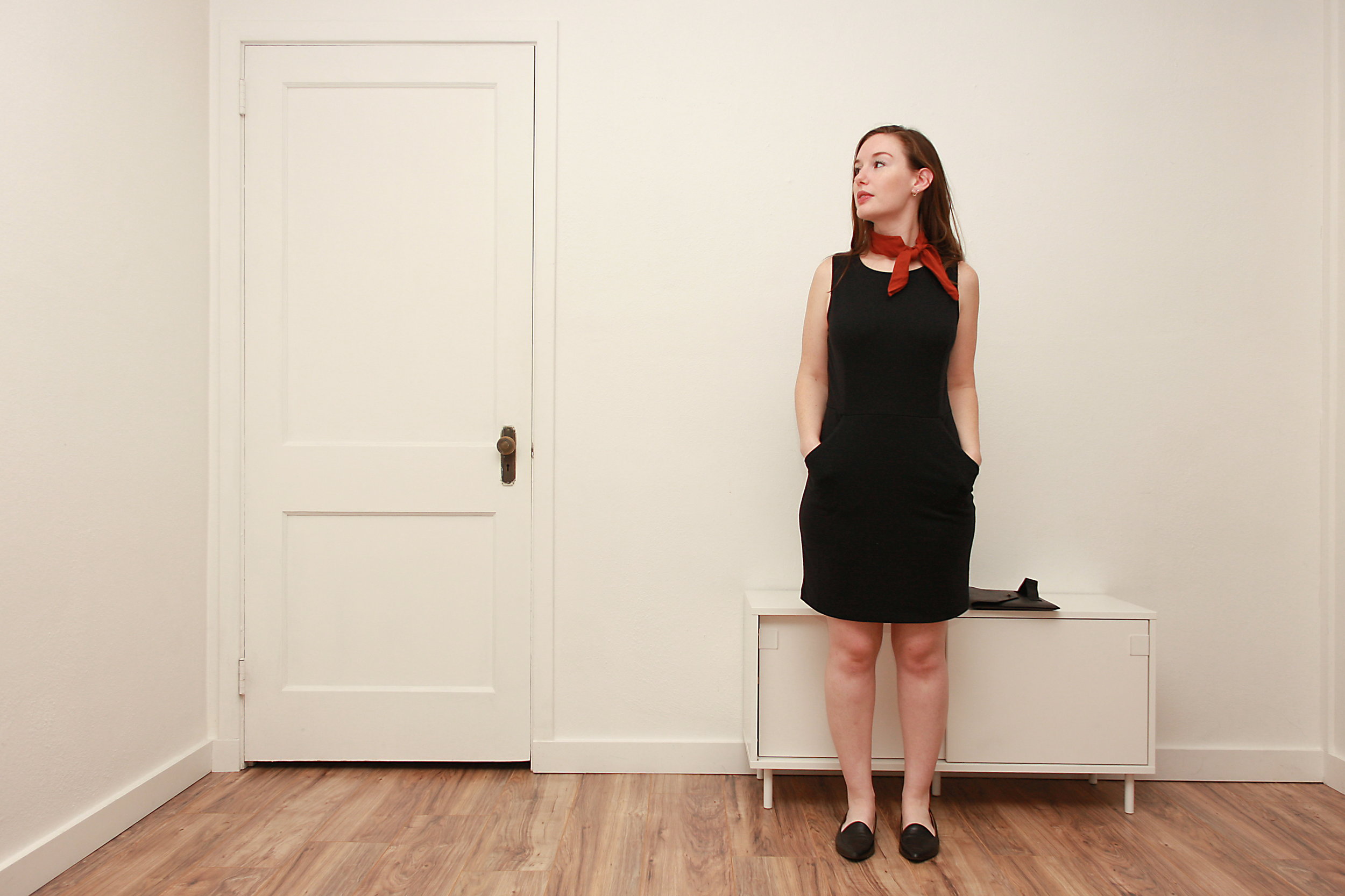 Alyssa wears a black sheath dress with black flats and a rust bandana and places her hands in her pockets