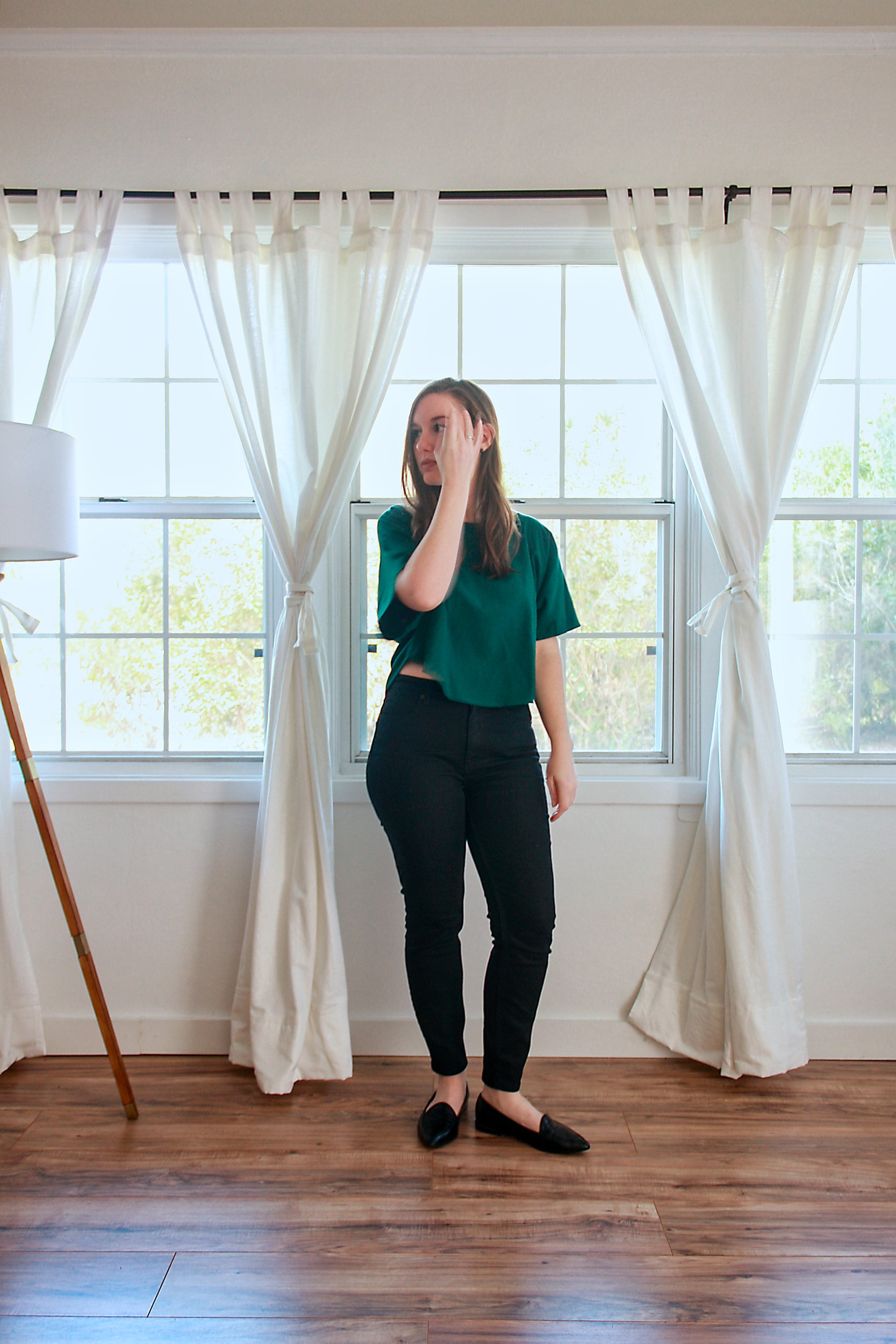 Alyssa wears a green top, black jeans, and black flats while brushing her hair from her face