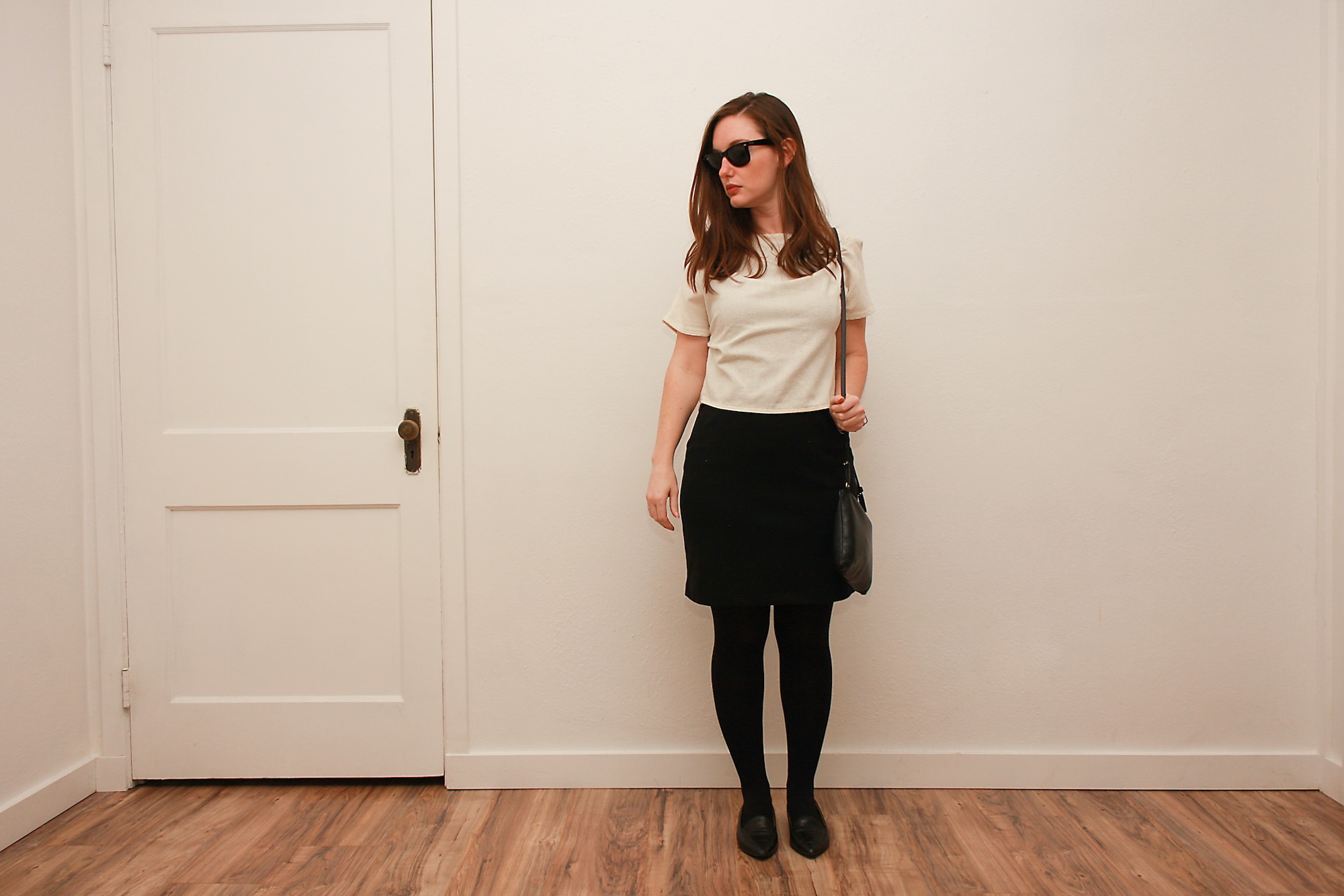 Alyssa wears a white silk tee over a black dress with tights and flats while holding a black purse