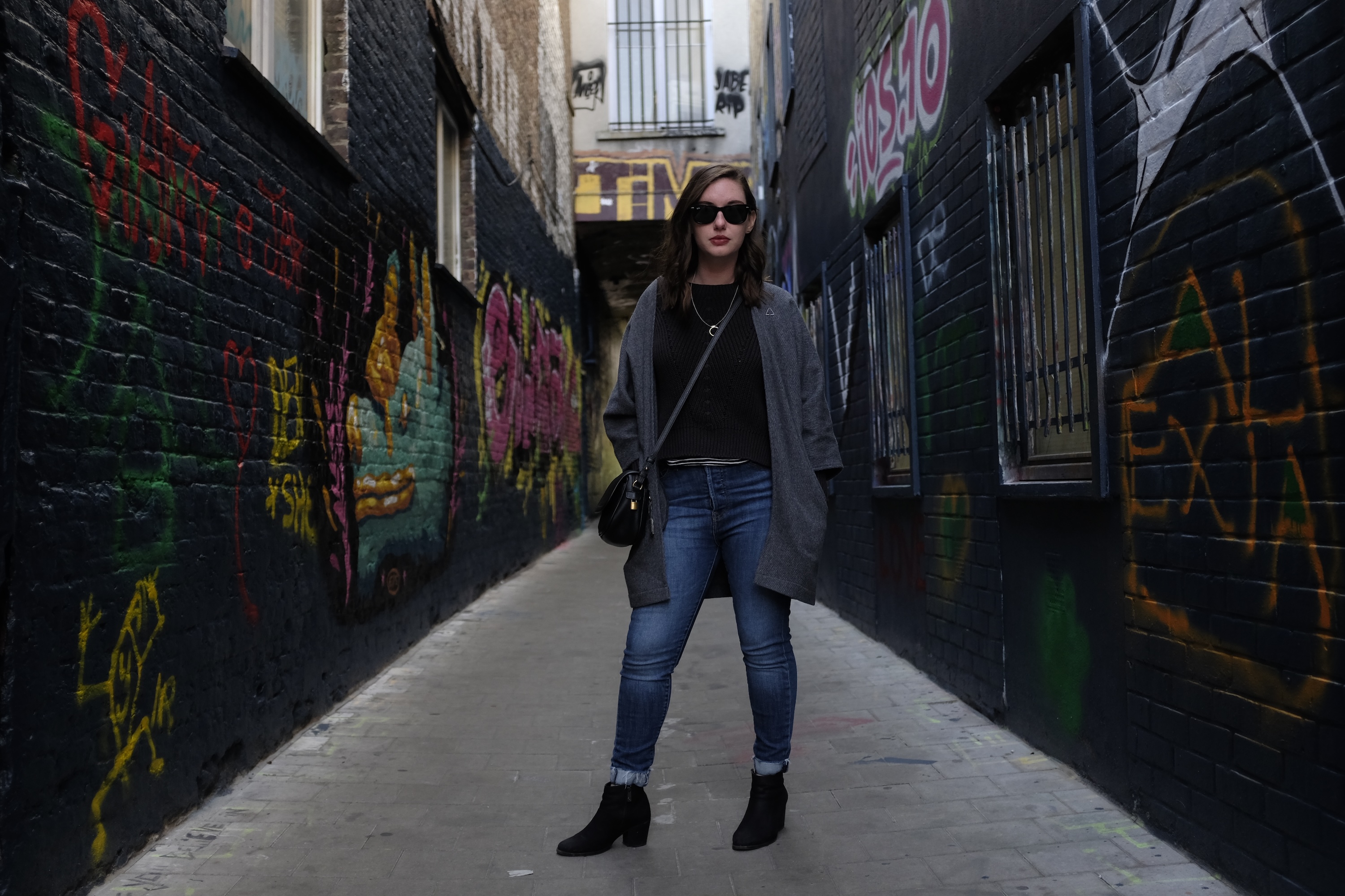 Alyssa wears blue jeans, a black top, black boots, and a sweater in the Ghent graffiti alley