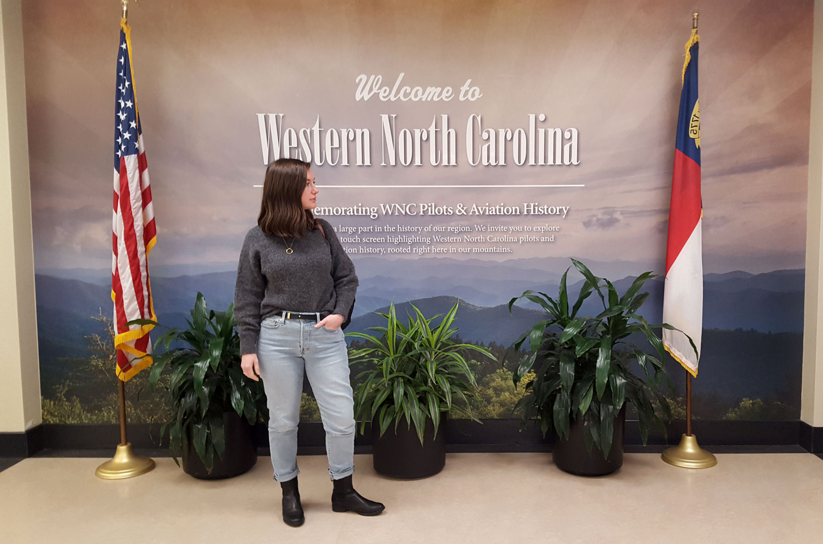 Alyssa stands in the Asheville airport wearing a sweater, jeans, and boots