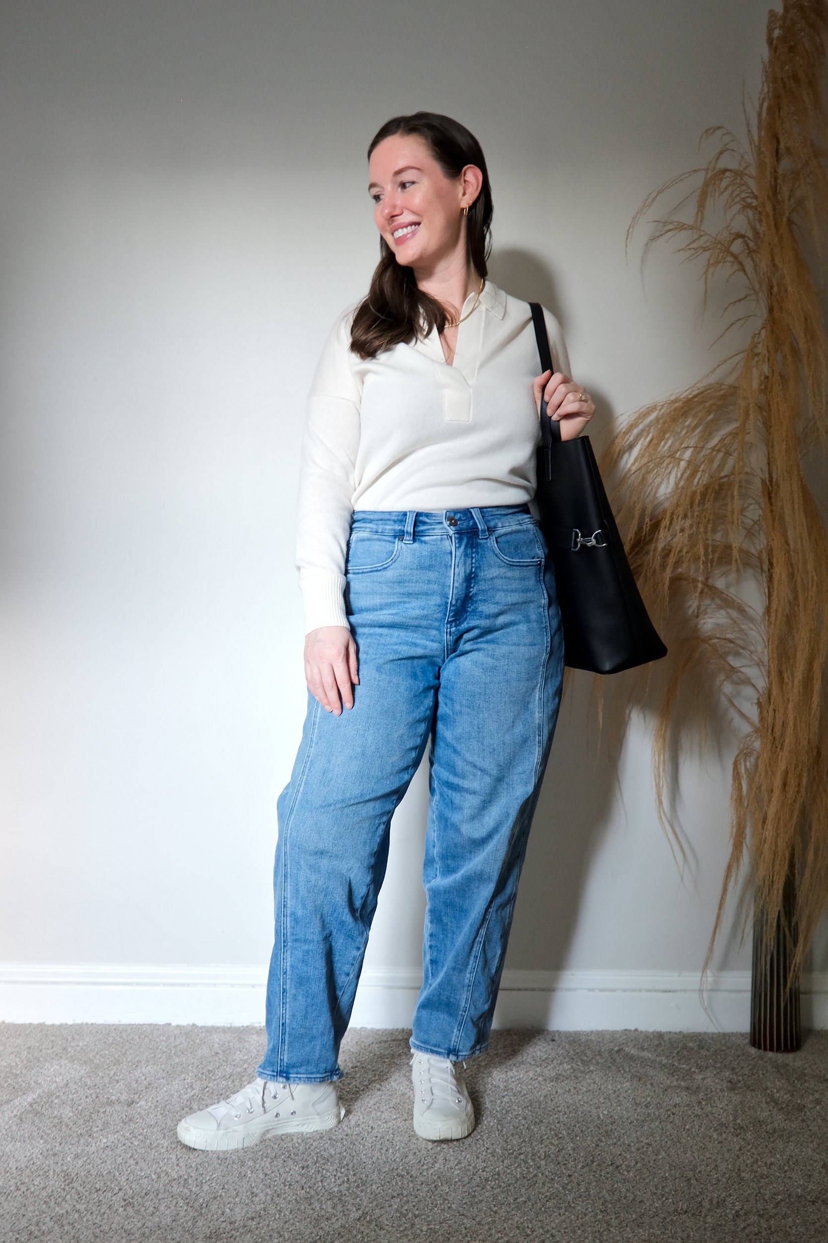 Alyssa wears the ivory cashmere polo sweater tucked into blue jeans with sneakers. She carries a tote and looks away from the camera.