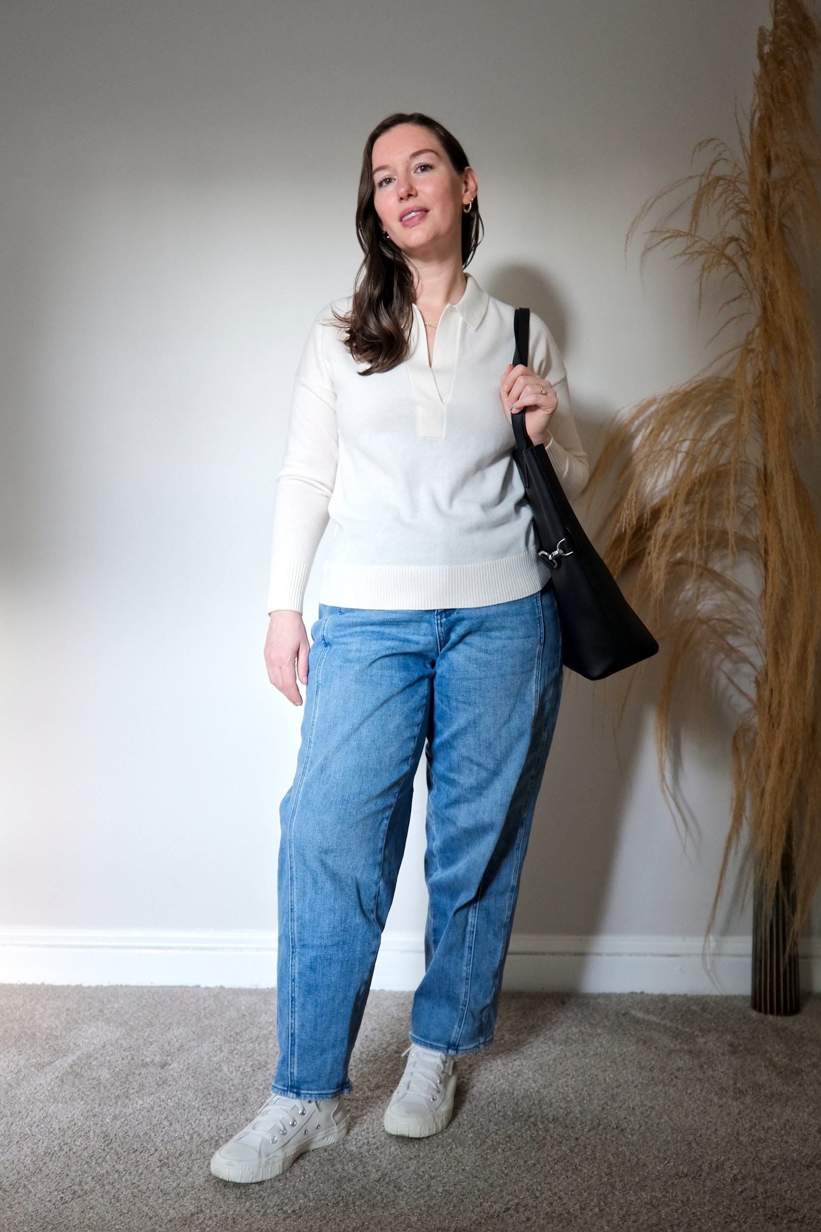 Alyssa wears the ivory cashmere polo sweater untucked with blue jeans and sneakers. She carries a tote and looks at the camera.