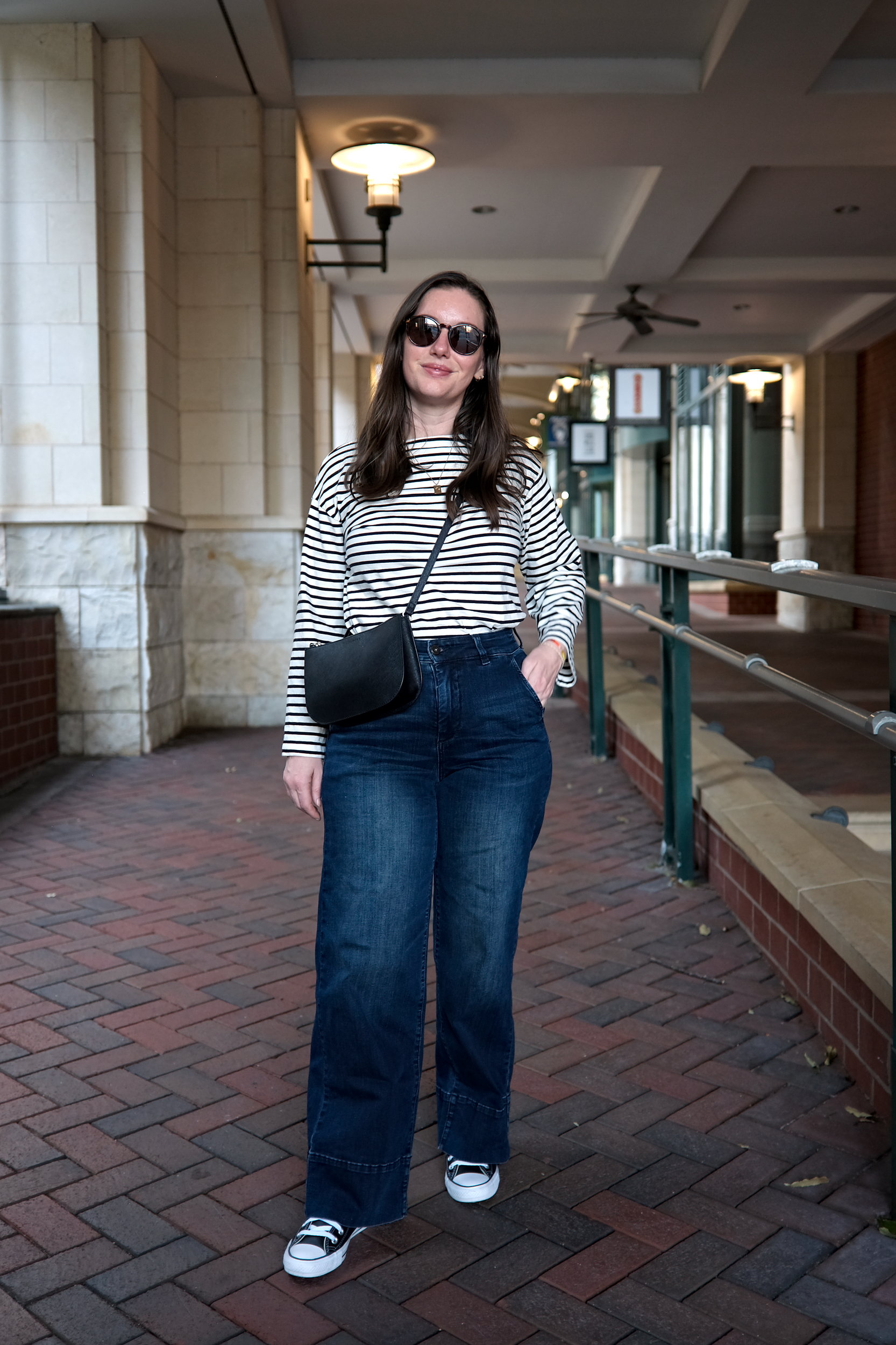 Alyssa wears the Carol High Rise High Stretch Jeans with a striped tee, sneakers, and sunglasses. She faces the camera and puts her hand in her pocket.