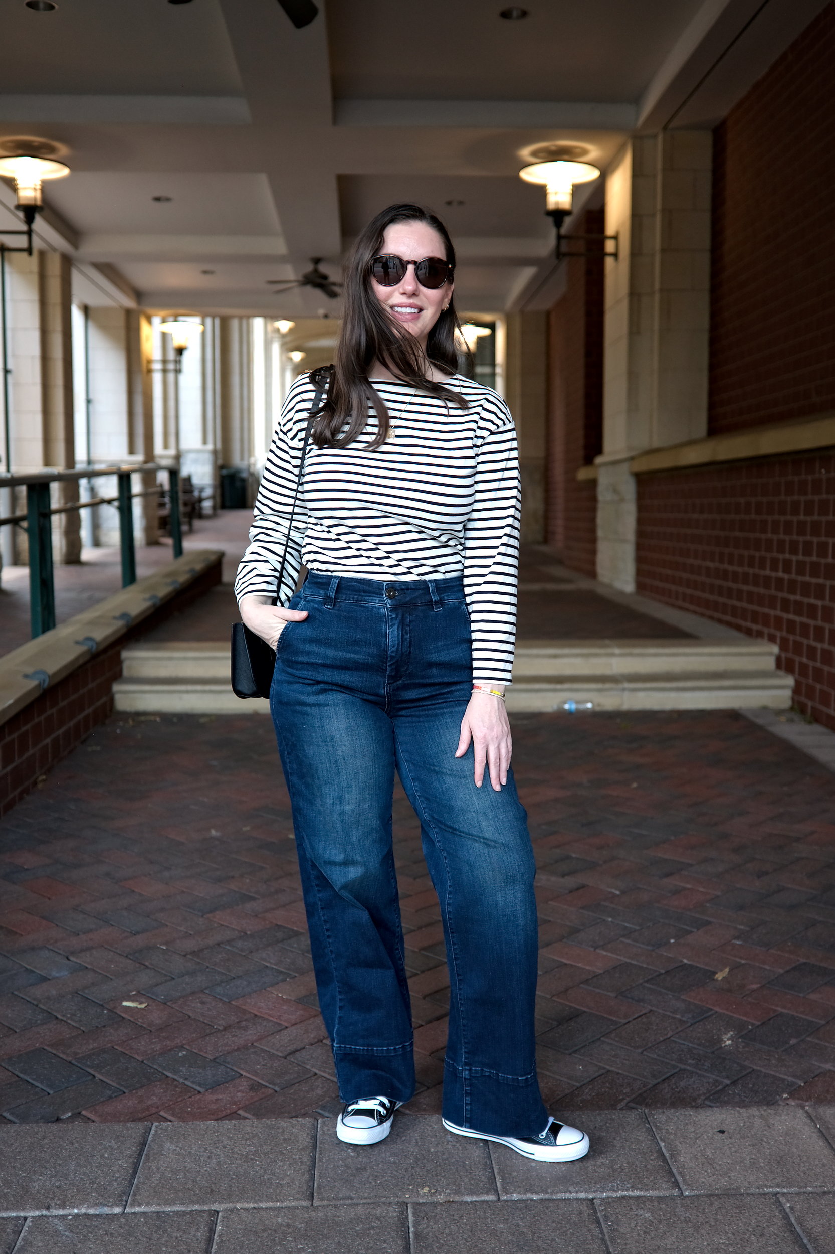 Alyssa wears the Carol High Rise High Stretch Jeans with a striped tee, sneakers, and sunglasses. She faces the camera and puts her hand in her pocket.