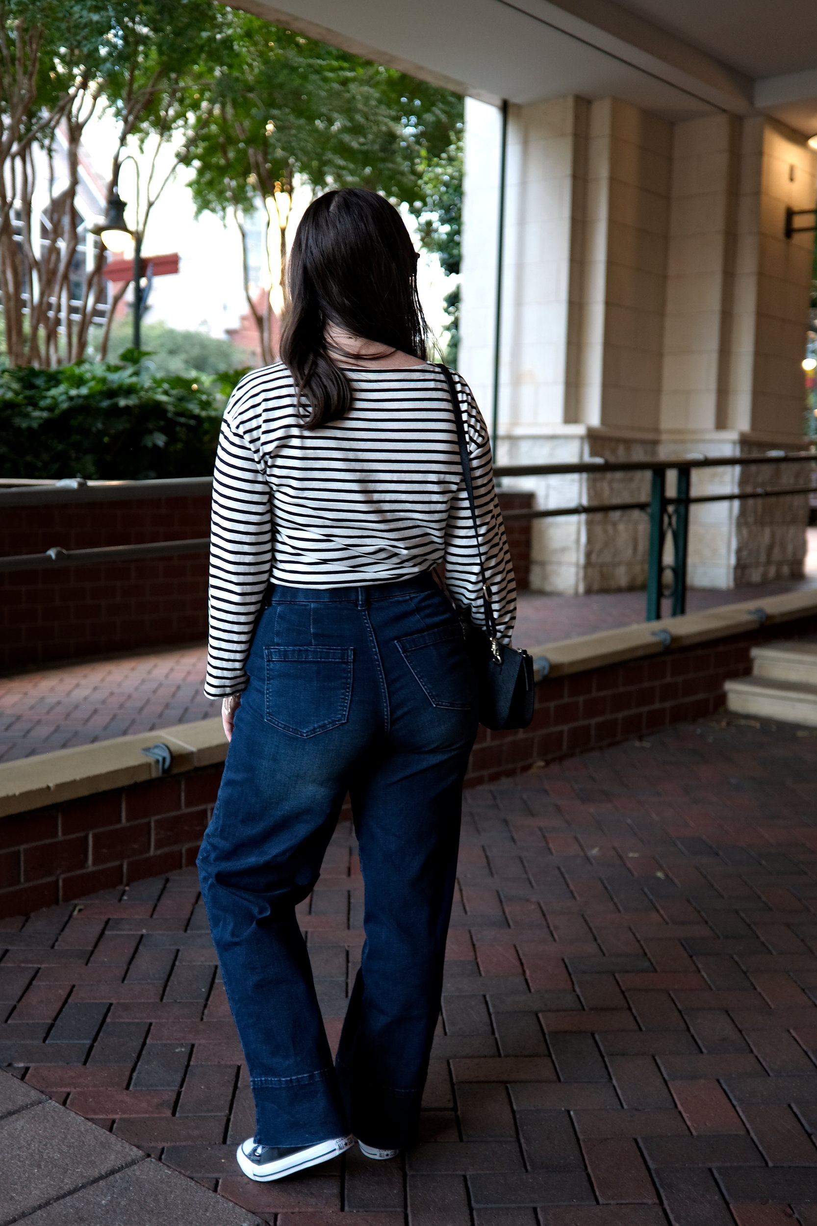 Alyssa wears the Carol High Rise High Stretch Jeans with a striped tee, sneakers, and sunglasses. She faces away from the camera.