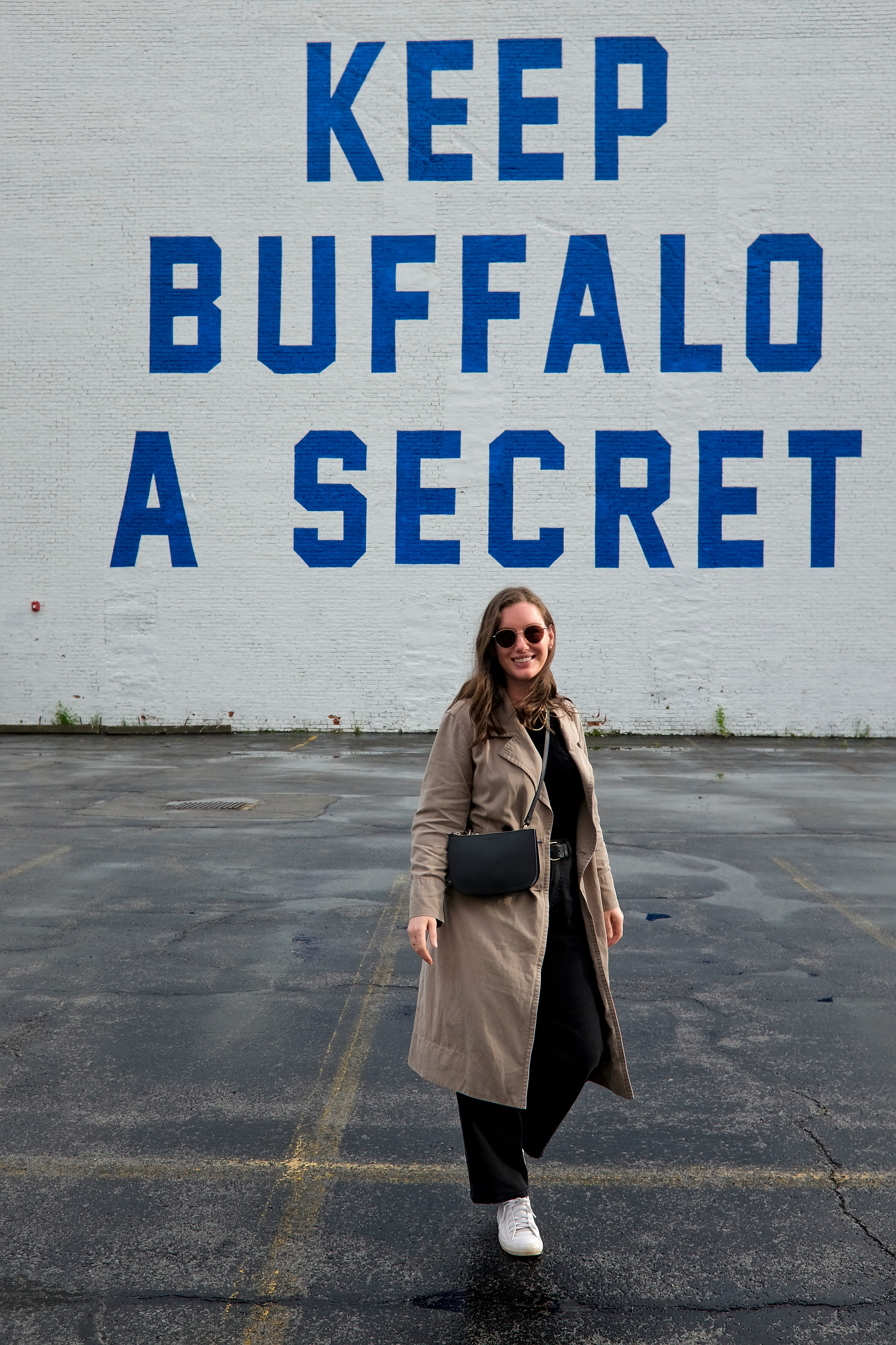 Alyssa walks in front of the Keep Buffalo a Secret mural in a black top, pants, and a trench