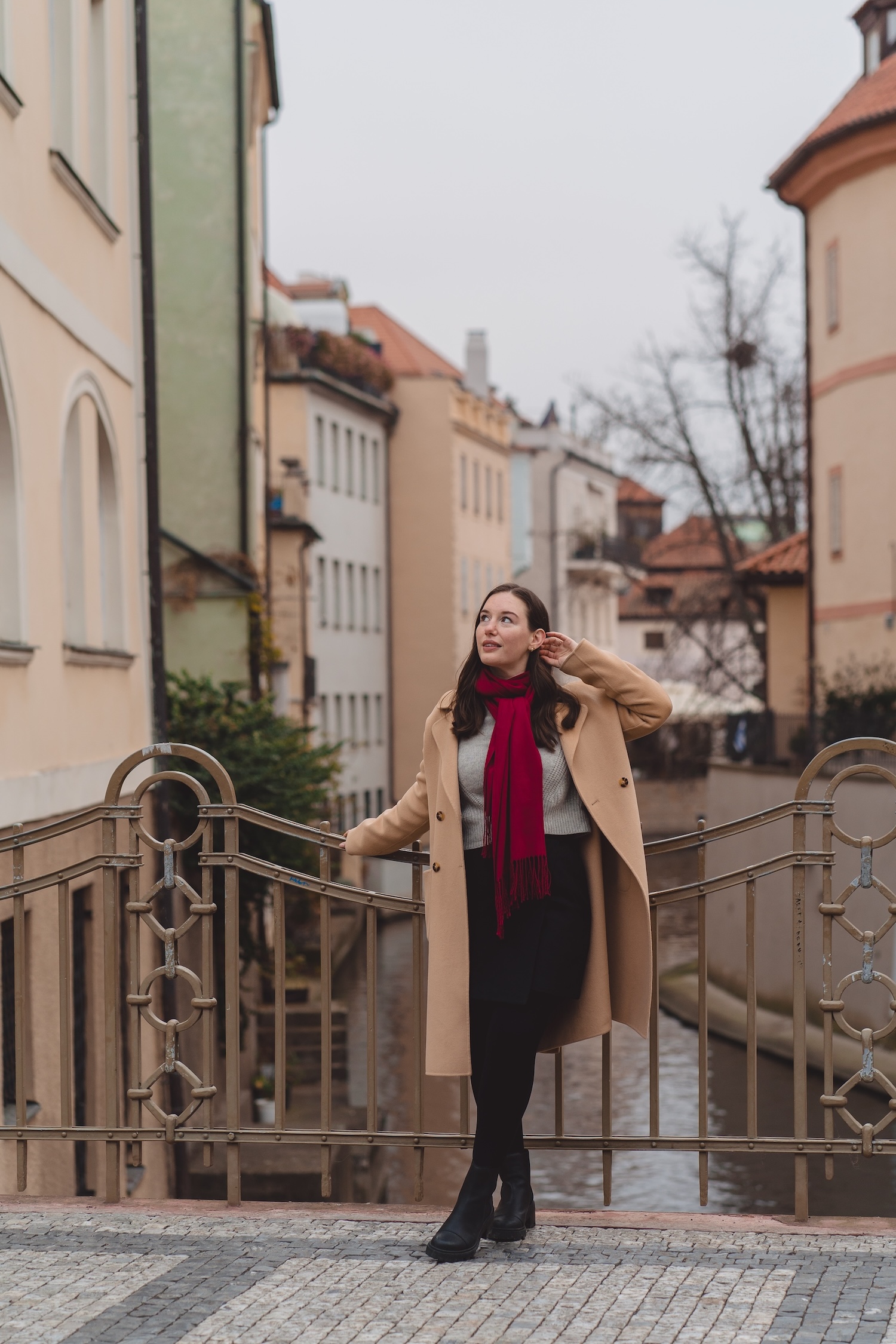 Alyssa wears a camel coat, skirt, and grey sweater on a bridge in Prague