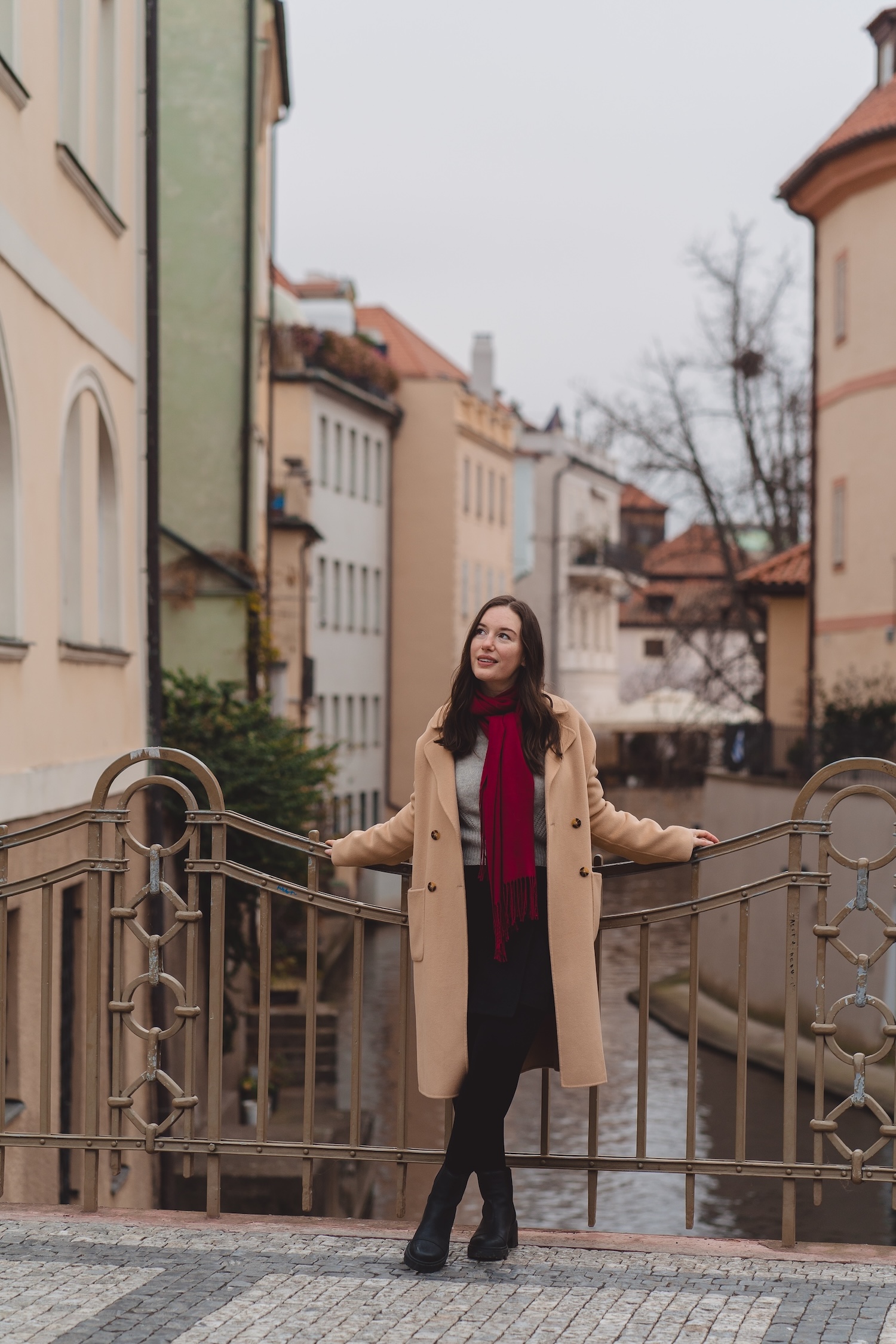 Alyssa wears a camel coat, skirt, and grey sweater in Prague