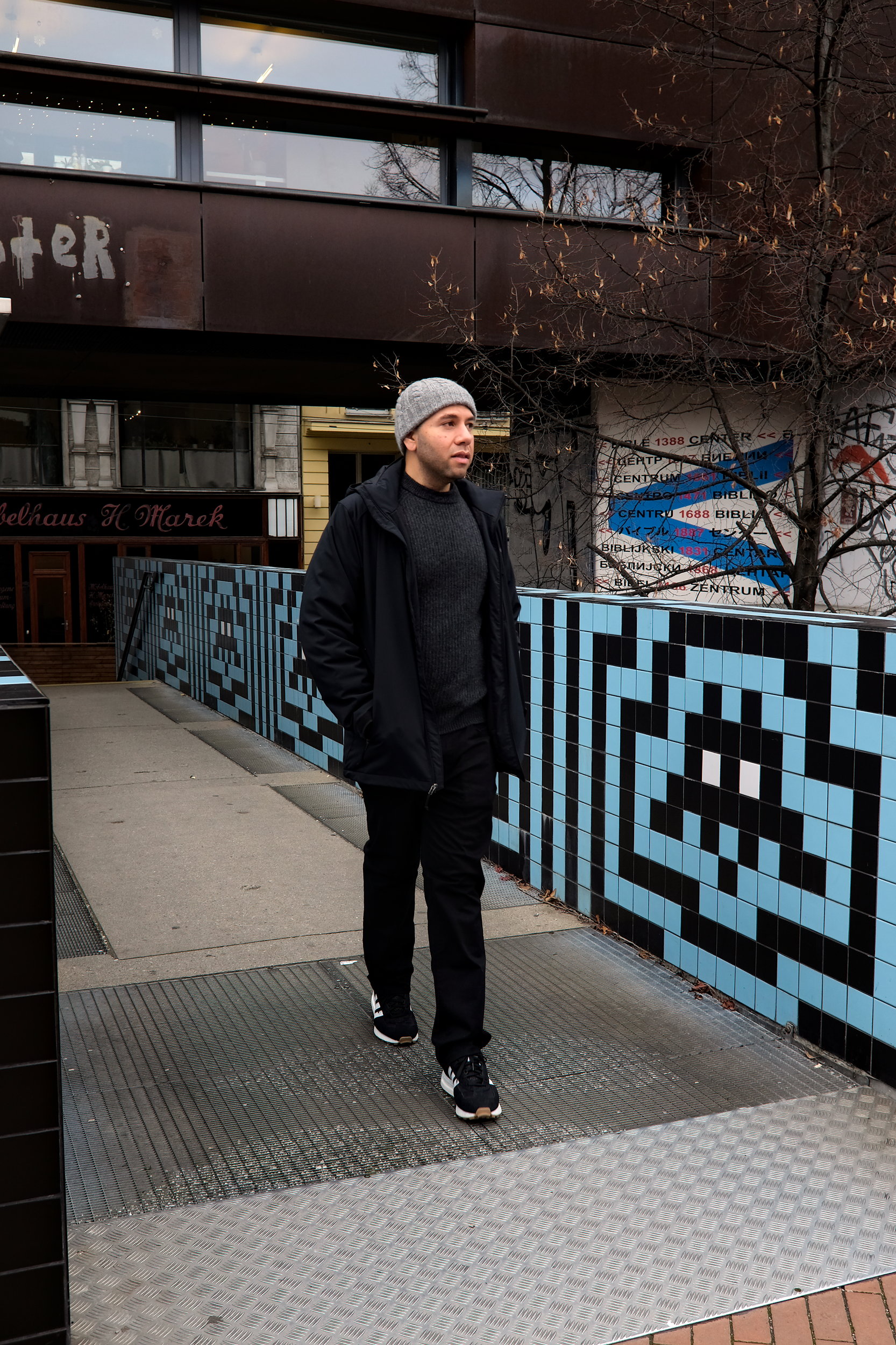 Michael walks on a bridge in Vienna, wearing black jeans, a grey sweater, a jacket, and a beanie